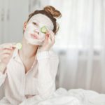 A cute girl with a face mask covers one eye with a slice of cucumber