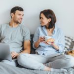 Couple sit together on bed, use laptop computer, have pleasant conversation between each other