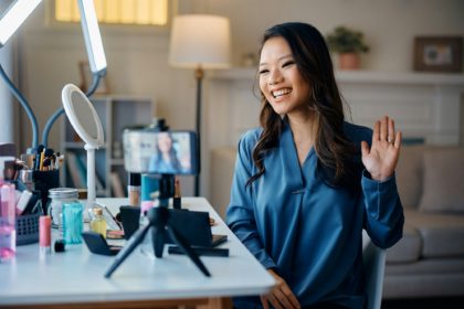 Happy Asian beauty influencer greeting her followers while recording makeup vlog at home.